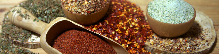 A wooden scoop and bowls filled with various colorful ground spices and herbs, including red chili flakes, green herbs, and a brownish-red powder, arranged on a wooden surface.