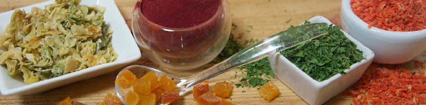 A variety of dried and powdered ingredients, including herbs, fruit pieces, and powders, displayed in small bowls and a clear spoon on a wooden surface.