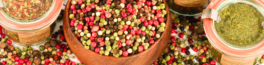 A wooden bowl filled with mixed peppercorns sits surrounded by glass jars of various dried herbs and spices, with some spices scattered on the surface.