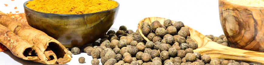 Bowls of powdered spices, cinnamon sticks, and whole black peppercorns in a wooden spoon are arranged on a white surface, highlighting various natural cooking ingredients.