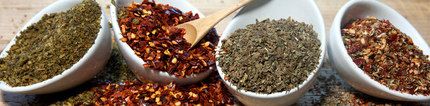 Four white bowls filled with different dried herbs and spices, including crushed red pepper flakes and green dried herbs, displayed on a wooden surface with a small wooden spoon in one bowl.