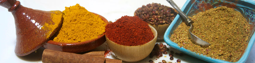 A variety of colorful spices including turmeric, paprika, and ground spice blends in bowls, with cinnamon sticks and a small pot on a white surface.