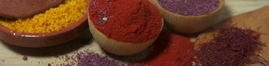 Close-up of bowls filled with vibrant yellow, red, and purple powdered spices, with some spilled onto a wooden surface alongside threads of saffron.