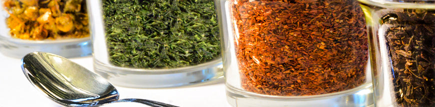 Close-up of glass jars filled with various dried herbs and spices in different colors, with a metal spoon placed in front of them on a white surface.