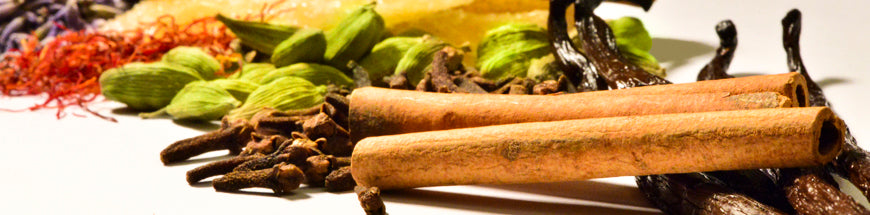 Close-up of assorted spices including cinnamon sticks, cloves, cardamom pods, vanilla beans, saffron threads, and other colorful spices arranged on a white surface.