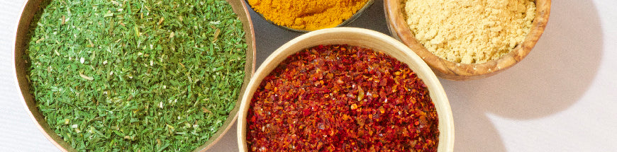 Four bowls filled with different spices: one with green dried herbs, one with red chili flakes, one with yellow turmeric, and one with light yellow ground ginger, arranged on a white surface.
