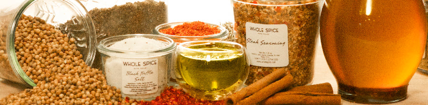 Various spices and seasonings in glass jars, ground and whole, are arranged on a table with cinnamon sticks, a small bowl of oil, and a bottle of honey or syrup.