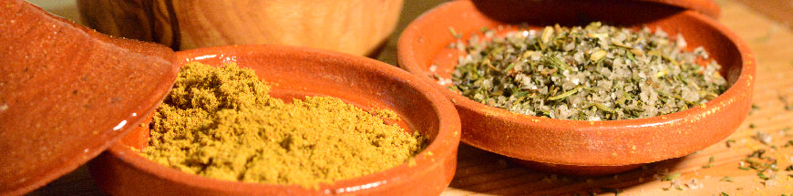 Two small terracotta bowls, one filled with ground yellow spice and the other with a greenish herb and salt blend, sit on a wooden surface with lids resting nearby.