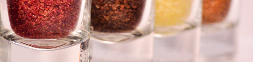 Close-up of four glass containers each filled with different colored salts: red, brown, yellow, and orange, arranged in a row on a light surface.