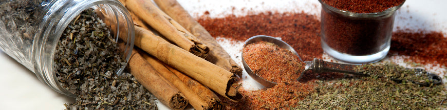 A glass jar of dried herbs, cinnamon sticks, a spoon with ground spices, a small glass of red spice, and piles of various loose dried herbs and spices spread on a white surface.
