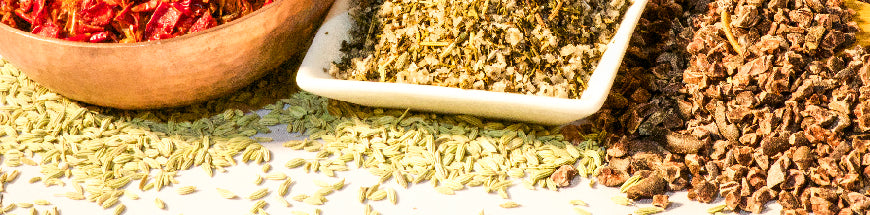 A close-up of assorted dried spices in bowls and spread on a white surface, including fennel seeds, crushed chili, dried herbs, and other blended spices.