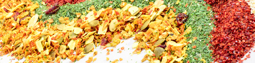 A close-up of colorful dried spices and herbs, including red flakes, green herbs, and yellow-orange pieces, arranged in wavy lines on a white surface.