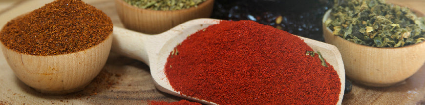 Wooden bowls and a scoop filled with various ground spices in different colors, including red, brown, and green, displayed on a wooden surface.