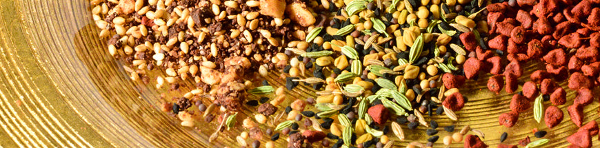 A close-up of assorted whole spices on a glass plate, including brown, green, and red seeds, arranged in colorful sections.