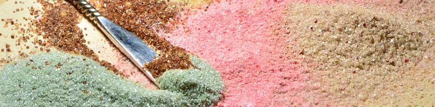 A close-up of piles of colorful sand—green, pink, tan, and brown—spread across a surface, with a small silver spoon resting on top.