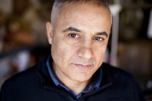 A middle-aged man with short gray hair and a serious expression looks directly at the camera. He is wearing a dark jacket, and the background is softly blurred.