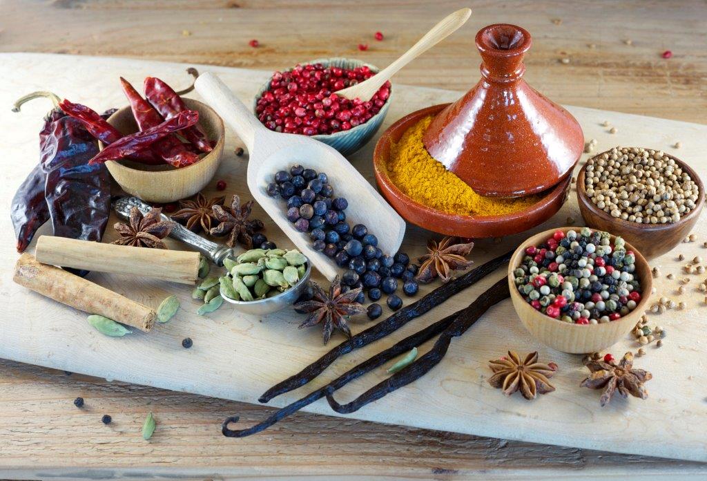 A variety of spices on a wooden board, including dried chilies, peppercorns, star anise, cardamom pods, juniper berries, coriander seeds, ground turmeric in a small ceramic tagine, and vanilla beans.