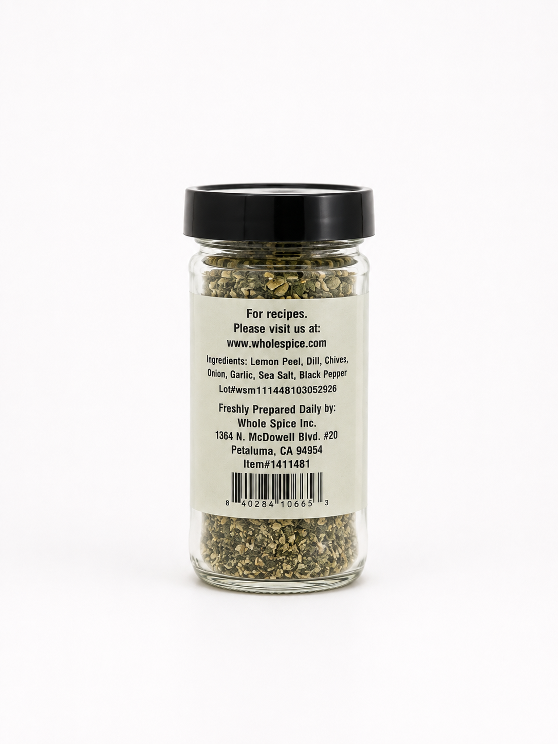 A glass spice jar with a black lid displays a label listing ingredients such as lemon peel, chili, and chives, along with contact details for the manufacturer, Whole Spice Inc. The jar contains Dill Seasoning—a mixed dried herb blend that serves as a versatile vegetable seasoning or a flavorful addition to chicken.