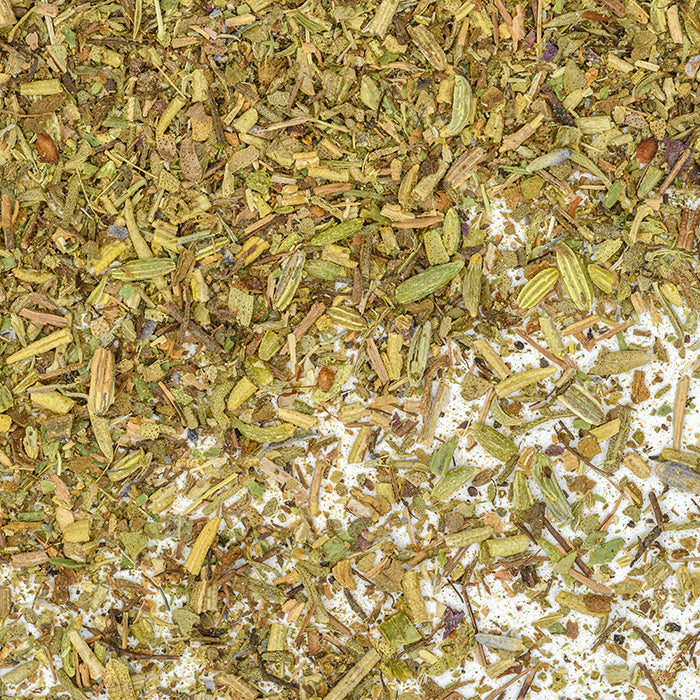 Close-up of Whole Spice Inc. Herbes de Provence Crushed, a salt-free French seasoning, showing small green and brown leaves and stems in a coarse blend with visible fragments on a white surface.