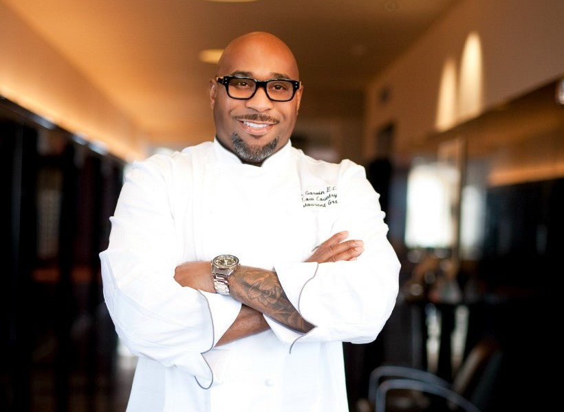 A smiling chef in a white coat and glasses stands with arms crossed in a well-lit hallway, wearing a watch and showing a tattoo on his forearm.