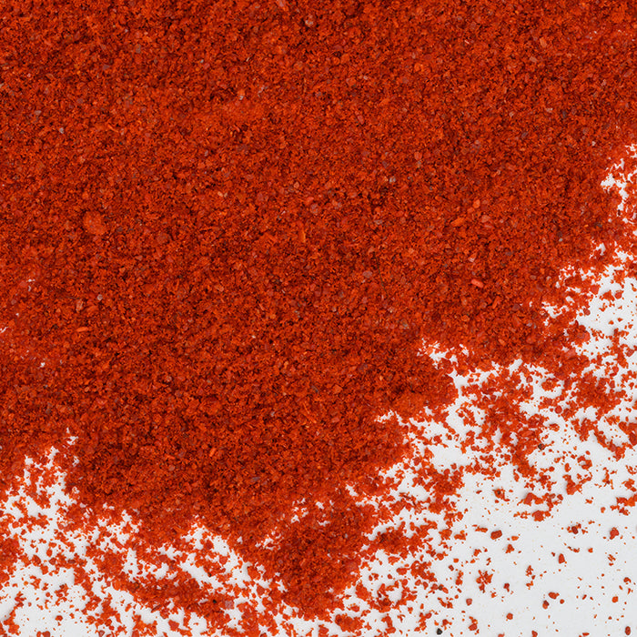 A close-up of vibrant California Paprika by Whole Spice, Inc. scattered on a white surface, with a bold concentration in the upper left and finer specks trailing to the lower right.