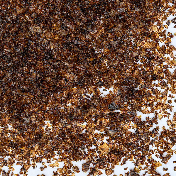 A close-up of Extra-Bold Smoked Sea Salt by Whole Spice Inc. shows dark brown flakes scattered on white, highlighting their irregular shapes and glistening texture.