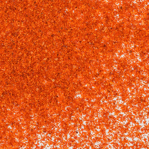 Whole Spice Inc. Harissa Spice, a bright orange-red chili blend, is scattered unevenly across a white surface, forming a vibrant textured pattern with some areas densely coated and others more lightly sprinkled.