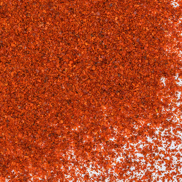 A close-up of Whole Spice Inc. New Mexico Chili Powder scattered on a white surface, showcasing its vibrant reddish-orange hue and coarse texture, perfect for adding heat and flavor to Mexican dishes.