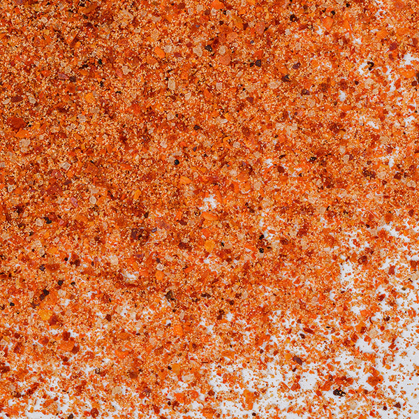 A close-up of Whole Spice Inc. Sriracha Hot Chili Seasoning shows its coarse, orange-red blend with varied textures and small flecks scattered across a white surface.