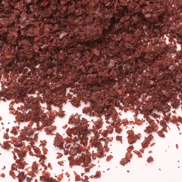 Close-up of Sumac Ground by Whole Spice Inc., with coarse, dark red flakes scattered on a white surface, highlighting their grainy texture and use as a tangy seasoning for various dishes.