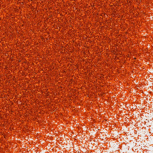 A close-up of Whole Spice Inc. Sweet Smoked Paprika scattered on a white surface, showing fine and slightly coarse orange-red granules clustered in the upper left and spreading to the lower right.