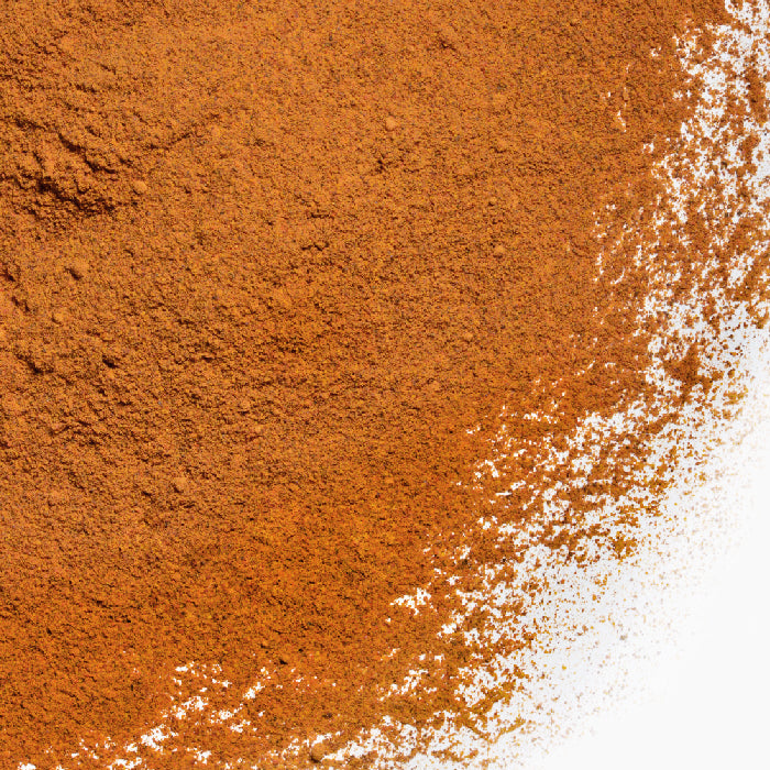 A close-up of Whole Spice Inc. Annatto Powder piled on a white surface, densely packed on the left and gradually scattering right, showcasing its vibrant color used for naturally coloring dishes.