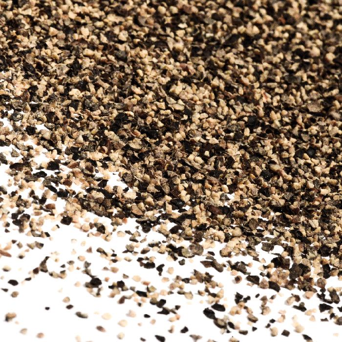 A close-up of Whole Spice Inc. Pepper Black Ground #2, called the King of Spices, scattered on a white surface, displays a blend of coarse and fine granules.