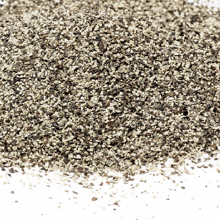 A close-up of Whole Spice Inc. Pepper Black Smoked Ground, the King of Spices, shows small, coarse black and tan specks scattered on a white surface.