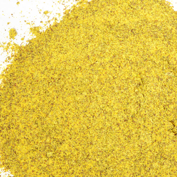 A close-up of Whole Spice Inc.'s Brown Mustard Seed Ground shows a fine, slightly granular yellowish-brown powder, unevenly spread on a white background.