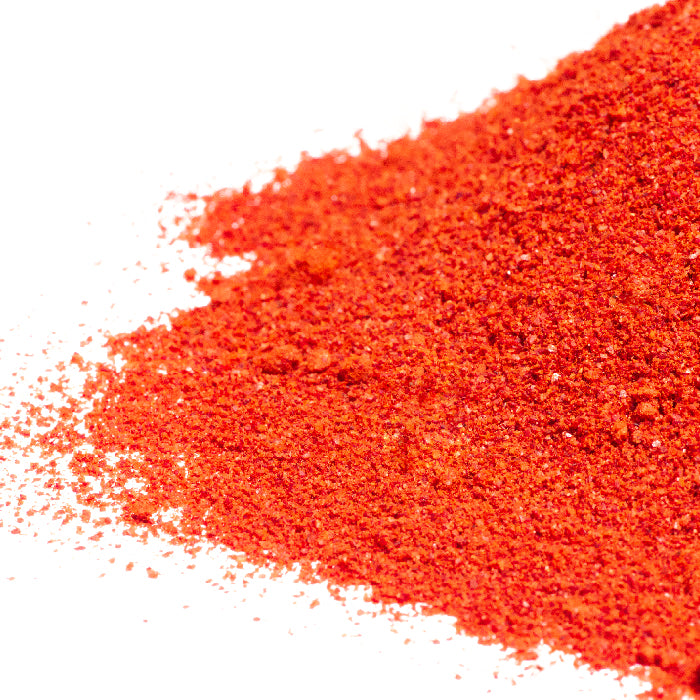 A close-up of Whole Spice Inc. California Chili Powder Organic, vibrant red-orange and scattered on a white surface—perfect for adding bold flavor to Mexican dishes.