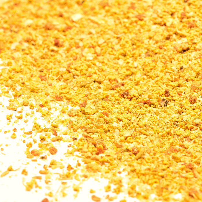 A close-up of Whole Spice Inc. Fennel Rub shows coarse, uneven yellow-orange granules scattered on a white surface—ideal for adding flavor and texture to roasted fish dishes.