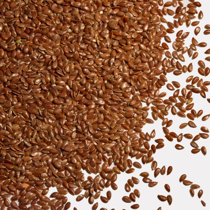 A generous heap of Whole Spice Inc. Flax Seeds is arranged on a white background, with a few seeds scattered loosely around the main pile.