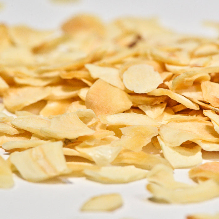 A close-up of Whole Spice Inc. Garlic Slices, showing dried, thin pieces scattered on a white surface.