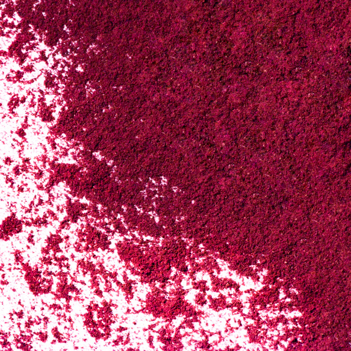 A close-up of Whole Spice Inc.'s Hibiscus Powder shows vibrant magenta powder, scattered across a white surface in dense and sparse areas, creating a dynamic, textured pattern reminiscent of crushed dried hibiscus.