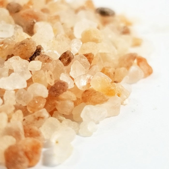 Close-up of Whole Spice Inc. Himalayan Pink Salt Coarse (3-5mm) crystals scattered on a white surface, displaying irregular shapes and natural shades of pink, orange, and white, naturally rich in trace minerals.