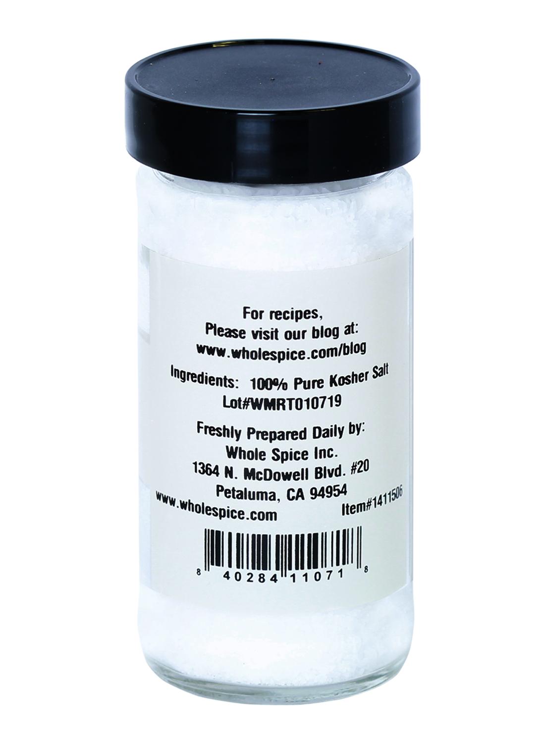 A clear plastic spice jar with a black lid from Whole Spice Inc. contains coarse white Kosher Salt flakes. The back label lists ingredients, lot number, manufacturer info, and a barcode. Enjoy its pure, crisp flavor in every dish.