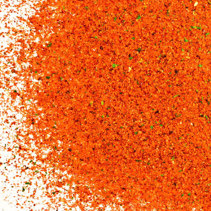 A close-up of Whole Spice Inc. Paella Spice Mix, vibrant orange-red with scattered green herb flakes, spread across a white surface.