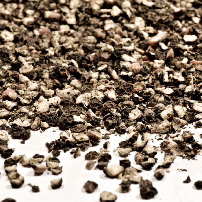 A close-up view of scattered dried and crushed Pepper Black Cracked by Whole Spice Inc., mingling with a hint of spice, on a white surface.