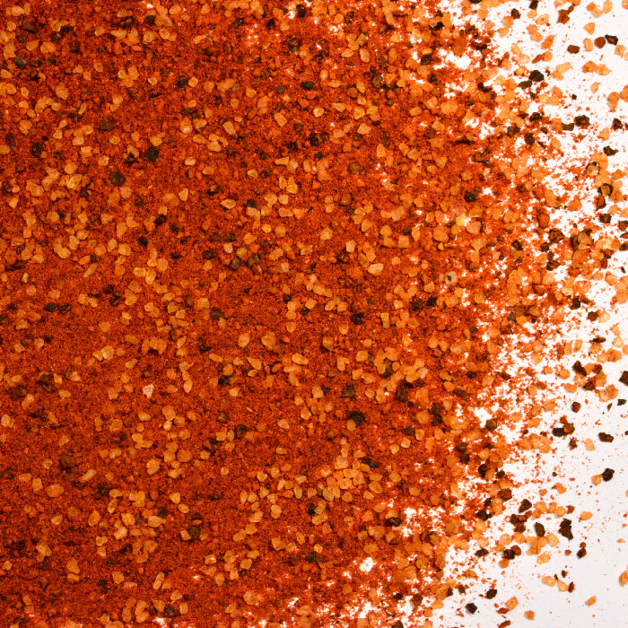 A close-up of coarse, red-orange Pica Pica Black Beans Seasoning by Whole Spice Inc., scattered on a white surface. Its grainy texture and vibrant specks add bold flavor to dishes like black bean salad.