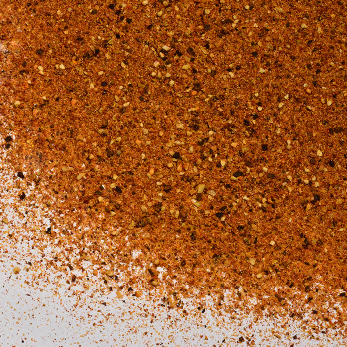 A close-up of Whole Spice Inc.'s Salt-Free Seasoning—a reddish-brown blend with coarse grains and bits, scattered on a white surface—ideal as a flavorful no salt seasoning.