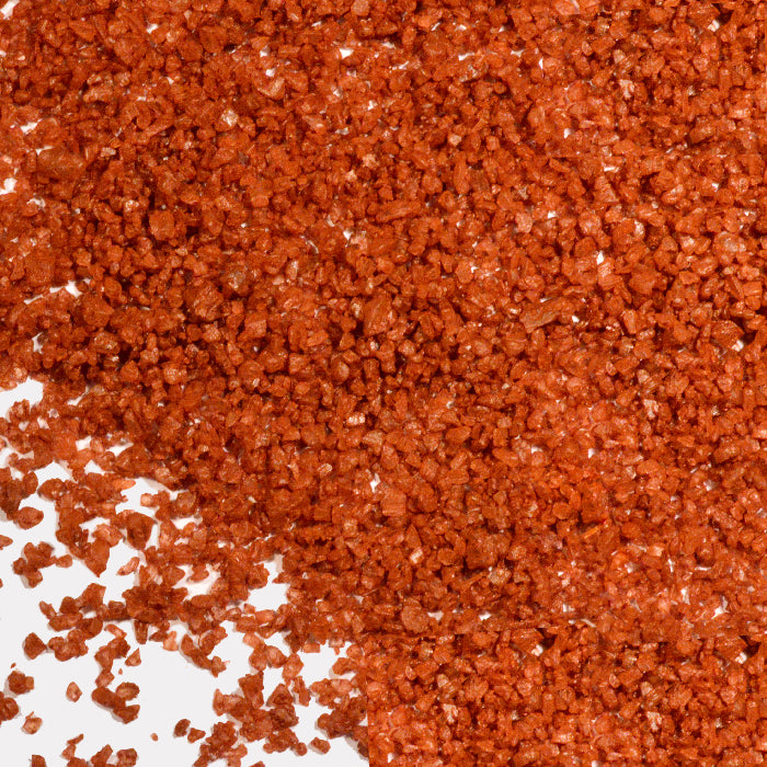 A close-up of coarse, reddish-orange granules scattered on a white surface, resembling Whole Spice Inc.’s Hawaiian Red Gold Sea Salt.