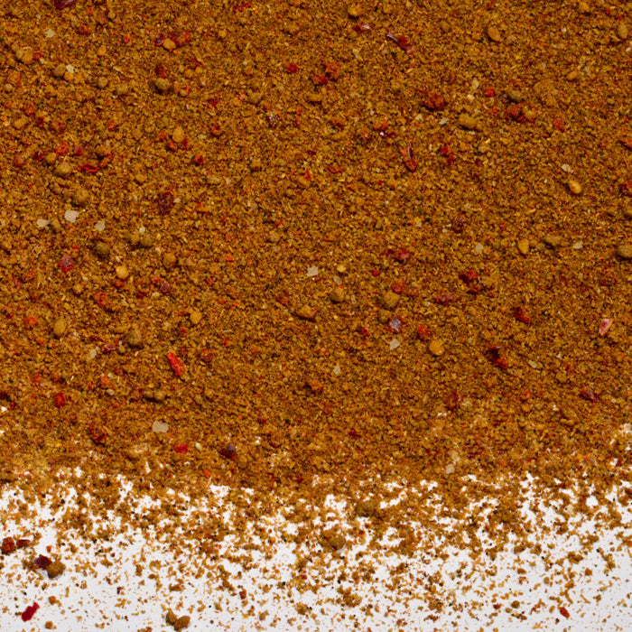 A close-up of Whole Spice Inc. Szechuan Seasoning—a vibrant reddish-brown blend with red, yellow, and tan flecks—scattered on a white surface, perfect for meat rubs.