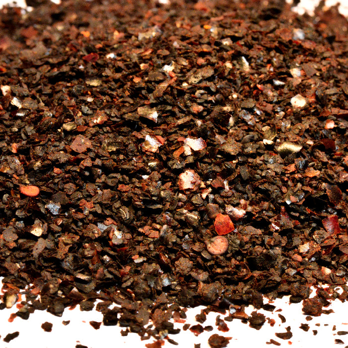 A close-up of Whole Spice Inc.'s Urfa Chili Crushed shows a pile of deep red, brown, and orange chili flakes with small white seeds scattered on a white background.