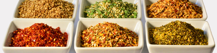 Six square white bowls filled with colorful spices and dried herbs, arranged in two rows. Each bowl contains a different blend, showcasing various textures and shades of yellow, green, orange, and red.
